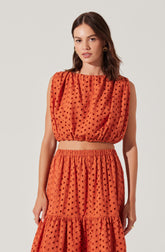 A woman is wearing a two-piece outfit made of orange fabric with black polka dots. The top is sleeveless with a gathered, slightly puffed shoulder design and an elastic waistband, creating a cropped look. The matching skirt is high-waisted, gathered at the elastic waist, with a tiered design that features a ruffled section toward the hem.